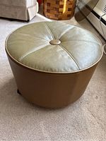 Side view of the round ottoman showing green vinyl top with stitching and a central button, tan vinyl sides, and caster wheels.