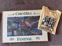 Framed 1992 Corn Hill Arts Festival poster alongside folded 2002 Corn Hill T-shirt on carpeted floor.