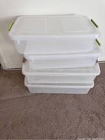 Four clear plastic underbed storage boxes stacked on carpet. Two smaller bins with green handles are visible.