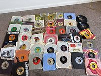 Photo showing a collection of 45 vintage vinyl 45 RPM records with assorted colorful paper sleeves from various record labels.