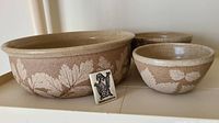 Three nested pottery bowls with speckled beige finish and white leaf motif, Wizard of Clay ceramic branding tile in front