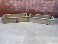 Photo showing two rectangular wooden window flower boxes placed on concrete with a brick wall background.