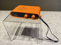 Front angled view of the orange Aitron transistor radio showing speaker grill on top, tuning knobs, and wrist strap.