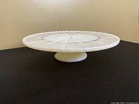 Cream white milk glass cake plate on pedestal shown from side angle on black surface with beige wall background.