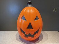 Frontal view of orange blow mold pumpkin jack o lantern with carved facial features, against dark background on light surface