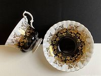 Teacup placed sideways next to saucer showing detailed gold floral pattern on black and white porcelain with gold trim.