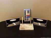 Overall group photo showing the Sheaffer calligraphy pen set in original packaging in the center, flanked by two gold pen/pencil gift sets in black velvet cases on either side. In front is the dual gold pen stand with agate stone base.