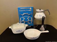 Vintage Corning Ware items including a teapot, metal cradle, casserole dish with lid, frying pan, and a detachable handle set against a neutral background.