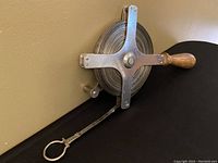 Side view showing metal reel with cross brace and wooden handle, metal tape extended with ring end.