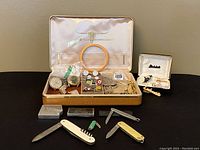 Full lot with vintage men's jewelry box open showing all items including rings, pins, bracelet, cufflinks, medals and assorted pocket knives arranged in front of it on black surface
