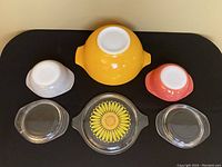 Three Pyrex bowls in orange, pink, and white with three corresponding clear glass lids, one with sunflower decoration