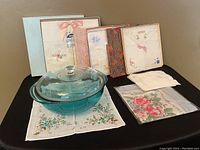 Photo showing clear teal Pyrex round casserole dish with lid placed on a floral linen napkin, several boxed sets of vintage decorative linen napkins behind it.