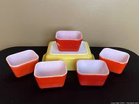 Five vintage Pyrex refrigerator dishes shown: four small red rectangular dishes and one large yellow rectangular dish beneath them, all with white interiors, displayed on beige surface.