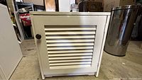 White wooden cabinet with louvered door viewed from front, showing single round knob and wear on surface.