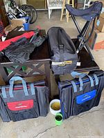 Camping gear items including folding tripod stool, two soft camping bleacher chairs in carrying cases, black sleeping roll on a table and ground.
