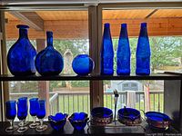 Wide display of all cobalt blue glassware items including vase, bottles, small vase or bowl, candle holders, goblets, creamer, sugar bowl and plates