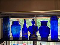 All six cobalt blue glass items on display in front of window: three jars with varying shapes and embossed designs, one vase, one soldier-shaped bottle, and one tall cylindrical jar with clamp lid