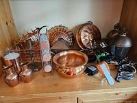 Shelf with copper items including two pitchers, a shallow bowl, lanterns, candle holders, clamp lights, copper plate with sun design, wire baskets, and folding wall hook
