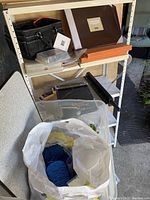 Crafting supplies on white shelf including paper cutter, scrapbooking albums, papers, scissor holder, plastic storage busting bag with yarn.