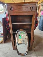 Full frontal and interior view of the wooden display cabinet and the oval wooden-framed mirror placed in front of it.