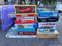 Stack of assorted board games and puzzle boxes in outdoor setting, showing multiple titles including Wit's End on top of the left stack.