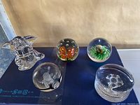Five glass paper weights including an elephant shape, two floral spheres, one with orange and one with red flower, one Pokémon character in a sphere, and a round clear paper weight with abstract embedded design on a blue surface.