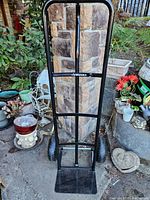 Front view of the black Husky hand cart/dolly with large 9.5 inch wheels, standing upright outdoors on paved surface near stone wall and garden plants.