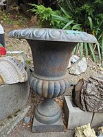 Full side view of large iron urn planter showing flared rim with leaf motif and bulbous section with vertical fluting, sitting on square base.