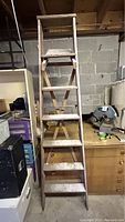 Front view of wooden ladder about 7 feet tall, showing six steps and small top platform. Located indoors against a concrete block wall with various items around.