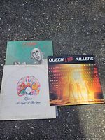 Three Queen vinyl records showing album covers for News of the World, A Night At The Opera, and Live Killers on a paved ground.