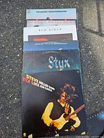 Seven vinyl records stacked on the ground showing titles from Steve Miller Band, Styx, Supertramp, Red Rider and The Raiders.