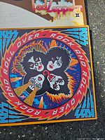 Photo showing Kiss vinyl record cover featuring stylized faces of band members surrounded by vibrant colors and text 'Roll Over Rock and Roll'