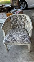 Front angled view of vintage rattan-style armchair with floral upholstered seat and interior backrest, showing weathered paint and wear.