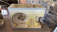 Shows the front of the original packaging featuring the Peter Rabbit bowl partially visible through a clear window, box surface has minor damage.