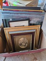 Box of multiple framed vintage art prints and wall art pieces stacked upright.