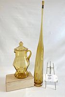 Photo showing amber glass jug with lid and tall amber vase against a white backdrop with a display stand.