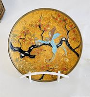 Brass decorative plate with cherry blossom tree and bird pattern on golden background, displayed on white stand.