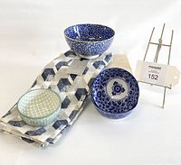 Four ceramic bowls arranged with a folded grey, blue, and white patterned cotton tea towel on a white surface. Includes a tag with lot number.