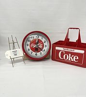 Photo of the metal Coca-Cola clock and red Coke bottle carrier side by side on white background.