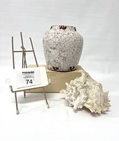White raku glazed pottery vase displayed on a wooden block, with metal stand and conch shell placed nearby.