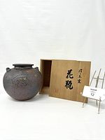 Full view of Shigaraki ware vase next to wood box with Japanese calligraphy and stand with lot number.