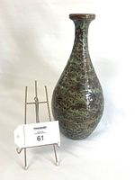 Front view of tall MCM ceramic vase with green and amber birdseye pattern, shown next to a small wire stand holding a maxsold tag.