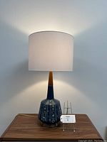 Full view of the lamp illuminated with white fabric shade, showing the navy blue chalkware body, teak neck and base, and brass harp.