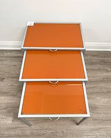 Three nested tables with orange tempered glass tops and silver metal frames