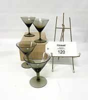 Four small smokey grey martini glasses arranged on and around a box, with a small easel holding the lot card