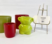 Four small succulent planters displayed including a red small cylindrical, red textured medium round, olive green rounded square, and light green bear-shaped planter along with a small easel holding a MaxSold lot card.