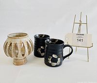 Lot showing two black ceramic mugs with cream decorative accents and a cream/tan ceramic tea light holder on a pedestal base, all photographed on white background with MaxSold tag.