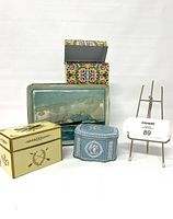 Front and top views of four vintage decorative tins arranged in a grouping, showing diverse patterns and colors including yellow, floral, blue with classical images, and a scenic maritime rectangular tin.