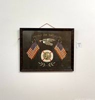 Framed vintage WWII silkwork memorial embroidery hanging on the wall showing full view including American flags, bald eagle, olive branches, and photo in lifesaver ring.