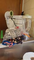 White Braun stand mixer with attached clear blender jug, beaters and whisks wrapped in plastic, and fabric aprons folded on top.